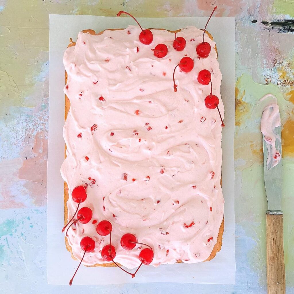 maraschino cherry chip cake. top down view of sheet cake uncut. cake is frosted with cherry frosting that is pink. upper right and lower left corners are garnished with stemmed maraschino cherries. background is abstract pinks, whites and tans. there is a wooden handled spatula with frosting on it positioned next to the cake. 