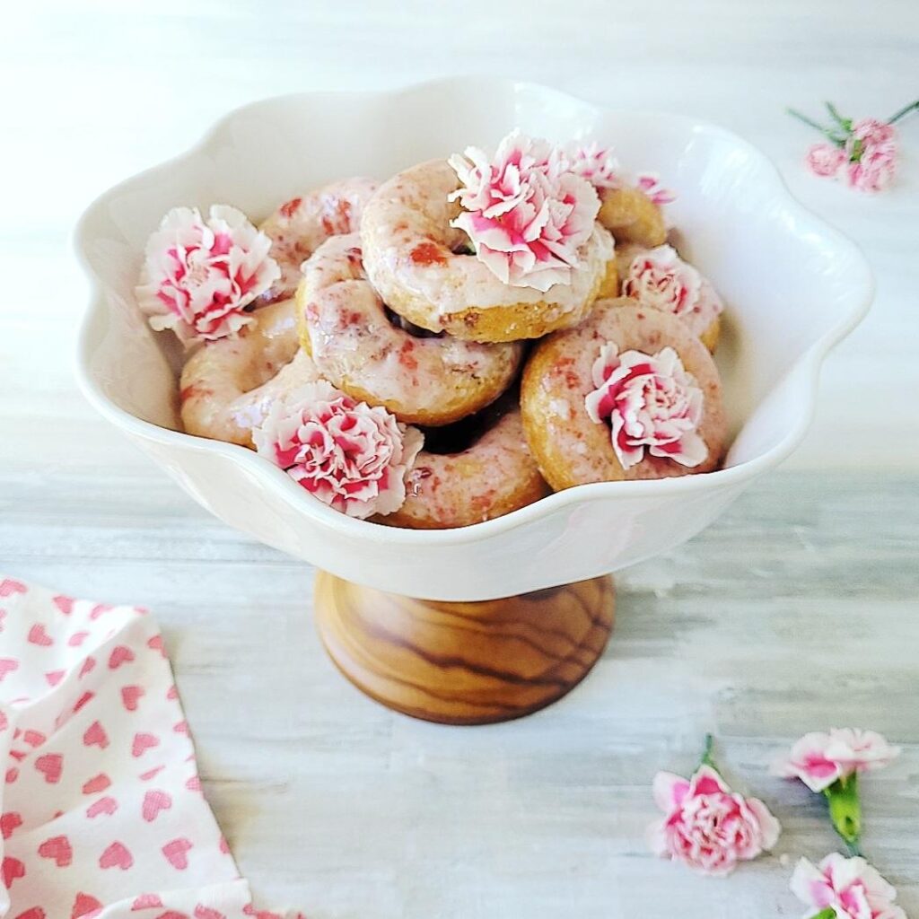 baked cherry donuts with pink cherry glaze and decorated with pink carnations are piled on a white ceramic cake stand with a wooden base. 