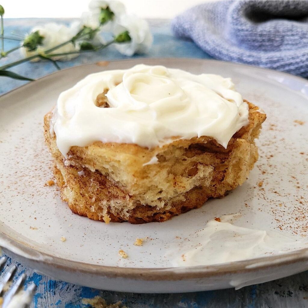 soft and fluffy cinnamon roll on a gray plate sprinkled with cinnamon. side view of the round cinnamon roll so you can see the soft, fluffy interior. top is covered with thick cream cheese icing. background has a cozy knit sweater and tiny white flowers. 