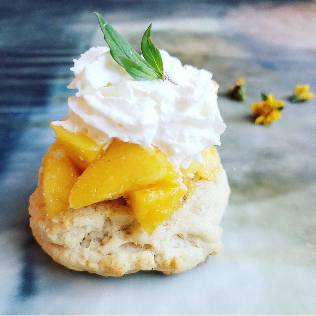 peach shortcake. side view of a homemade biscuit piled with honeyed peaches and topped with whipped cream and a sprig of mint. there are tiny yellow flowers in the background. 