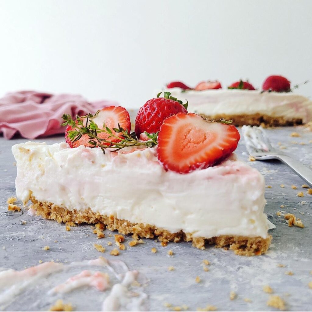 no bake strawberry swirl cheesecake with pretzel crust. close up side view of one slice garnished with thyme and fresh strawberries. the remaining uncut cheesecake is in the back right corner. 