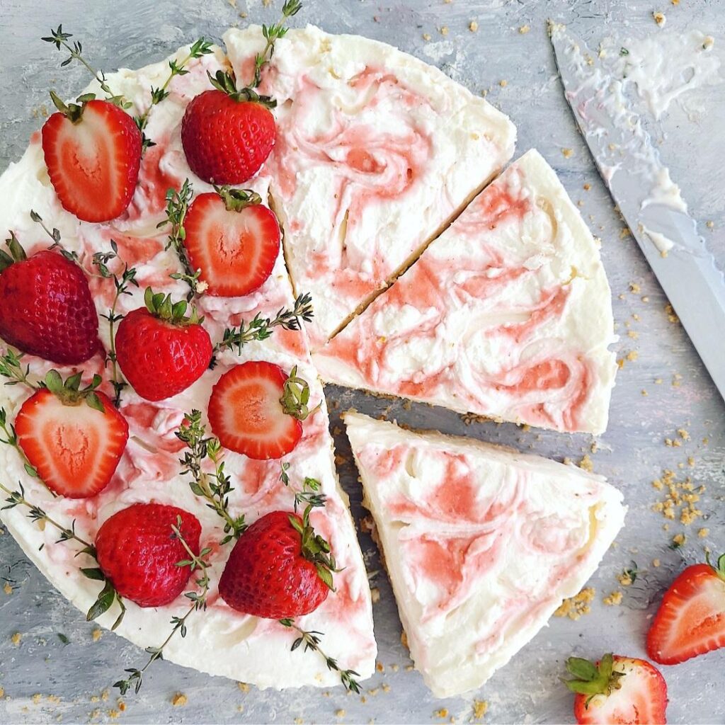 no bake strawberry swirl cheesecake. top down view of cake with three slices cut. the remaining uncut side of the cake is topped with fresh strawberries and sprigs of thyme. background is gray and styled with a cheesecake-covered knife blade, pretzel crust crumbs and a sliced strawberry.