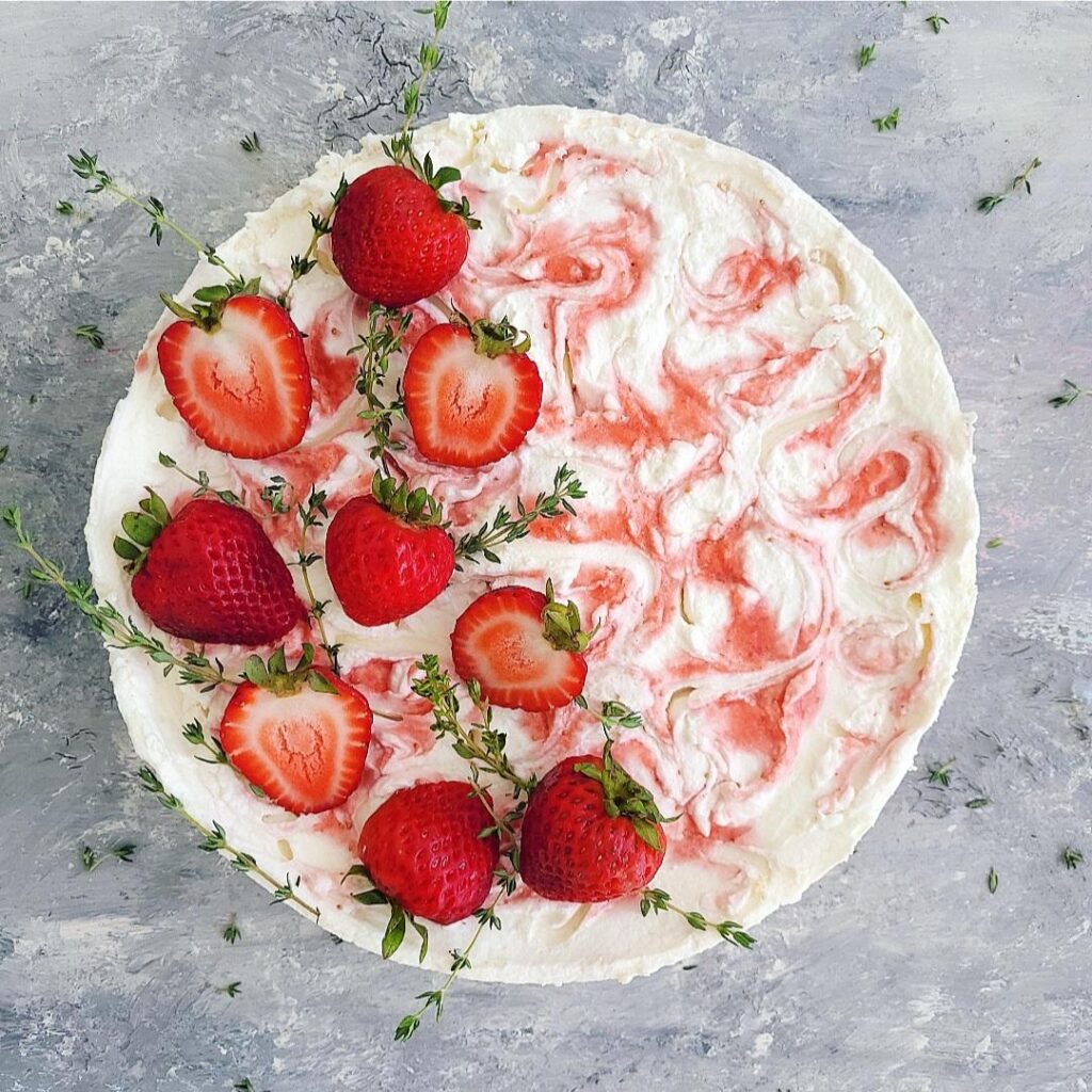 no bake strawberry swirl cheesecake. zoomed in top down view of uncut cheesecake. left half of cheesecake is garnished with fresh strawberries -- some are sliced in half and others are whole -- as well as sprigs of thyme. 