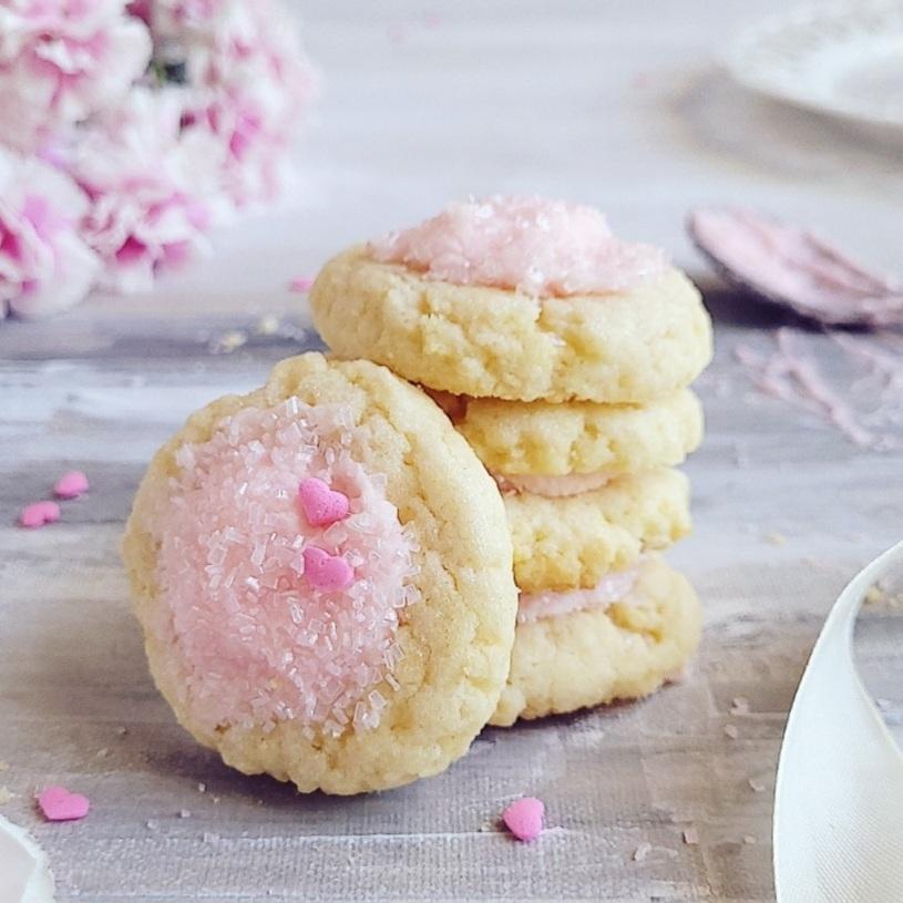 thumbprint cookies with icing. side view of buttery cookies stacked four high and frosted with pink buttercream. cookies are decorated with clear sprinkles and tiny pink hear candies. there are pink flowers in the back left corner. 