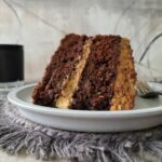 two layer chocolate banana cake with peanut butter frosting. slice of cake on a pale gray plate sitting on a contrasting gray fringed charger. background is gray abstract and there is a black candle in the back left corner.