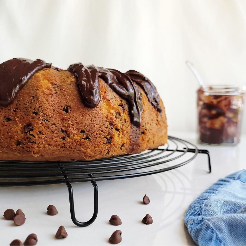 chocolate chip bundt cake with chocolate glaze. close up side view of uncut cake on a round baking rack. background is white and there is a glass jar of chocolate glaze in the back right corner. there are some loose chocolate chips in the front of the frame.