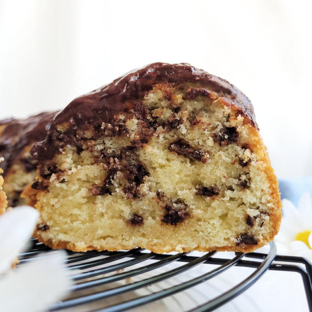 chocolate chip bundt cake. close up side view of one cut slice on a black wire baking rack. cake is topped with chocolate glaze. background is white. 