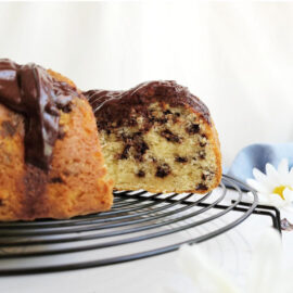 chocolate chip bundt cake. side view of cake on a black wire baking rack that is round. background is white. cake has one slice cut away so you can see the cake crumb and chocolate chips inside. cake is topped with a chocolate glaze.