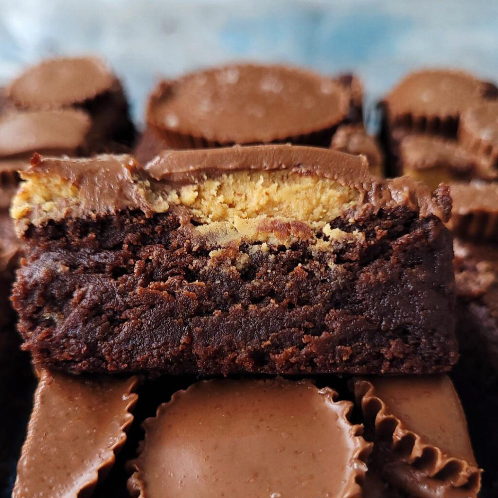 reese's peanut butter cup brownies. close up side view of a cut brownies so you can see the fudgy brownie layer and the peanut butter cup topping. background is blue. brownie slice is resting atop another slice.