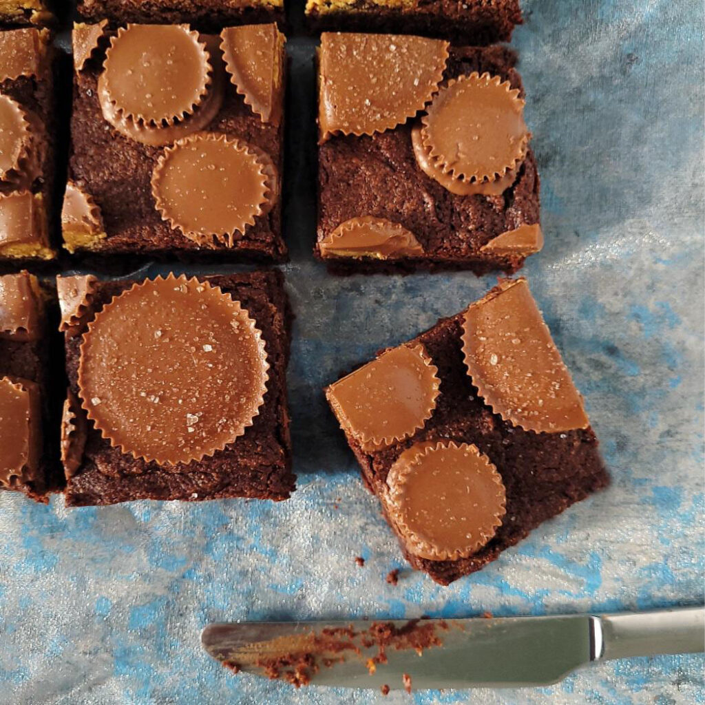 reese's peanut butter cup brownies. top down view of brownies topped with whole peanut butter cups (mini and king size). brownies are cut into squares and the bottom right slice is askew. background is gray with hints of vibrant blue. 