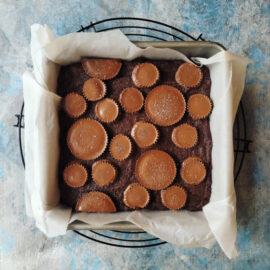 reese's peanut butter cup brownies. top down view of brownies topped with mini and king size peanut butter cups. brownies are uncut in an 8x8 baking pan lined with white parchment paper. baking pan is sitting on a round wire rack. background is abstract blue and white.