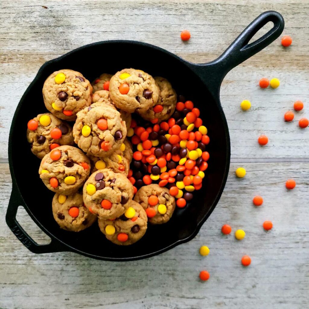 reese's pieces cookies with chocolate chips piled in a black cast iron skillet with loose reese's pieces around the cookies. top down photo. background is distressed wood. 