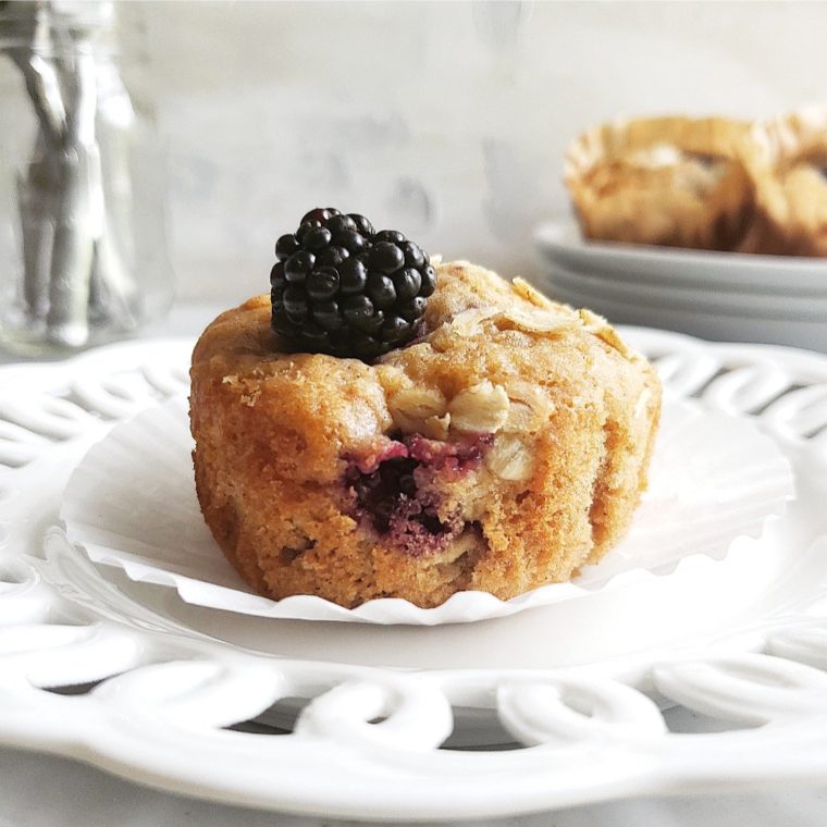 blackberry oat muffins. photo of a single muffin on a white plate with the muffin wrapper removed. muffin is topped with a fresh blackberry. 