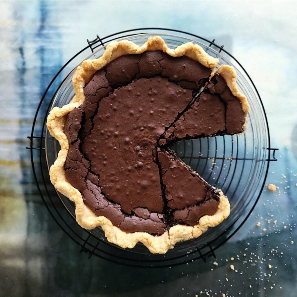 chocolate chess pie. top down view of pie in a glass pie plate on a round wire baking rack. two slices are cut and a third slice is missing leaving a void of pie crust crumbs. 
