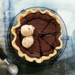 chocolate chess pie. top down view of pie in a glass pie dish on a black round wire rack. three slices are cut. the uncut portion has two scoops of coffee ice cream on top.