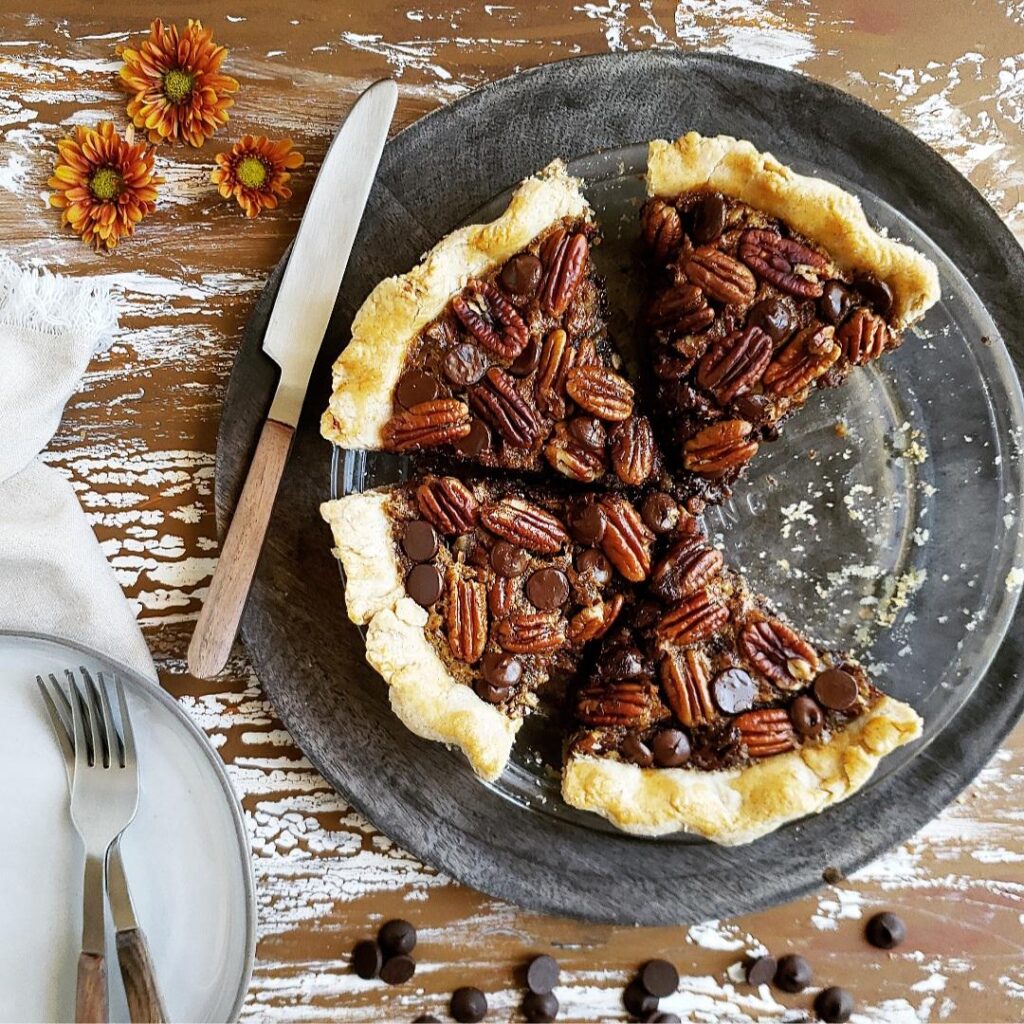 chocolate chip pecan pie in a glass baking dish. top down view of cut pie on a gray wooden charger. background is distressed brown wood with chipped white paint. image is styled with fall flowers a knife, forks and a plate.