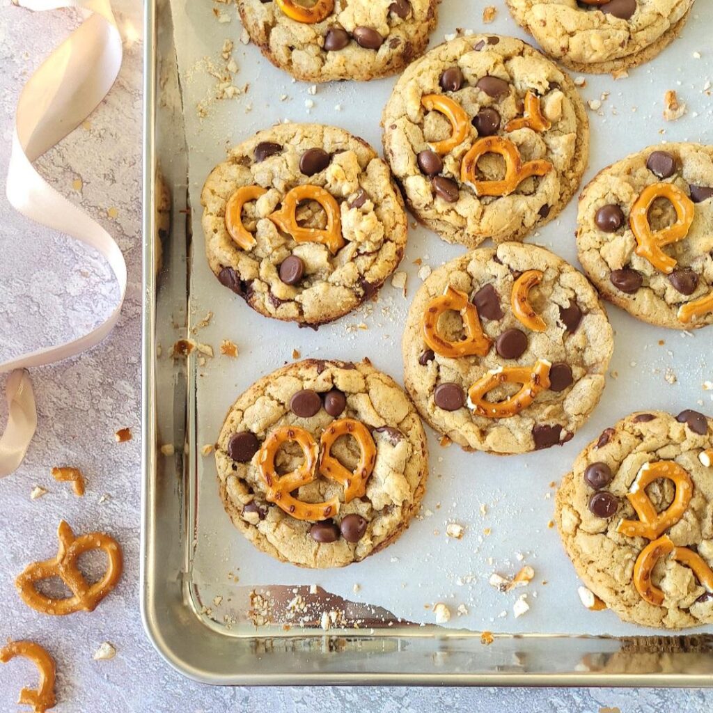 chocolate chip pretzel cookies on a stainless steel baking tray. top down view of baked cookies on white parchment paper surrounded by broken pretzel bits and crumbs. 