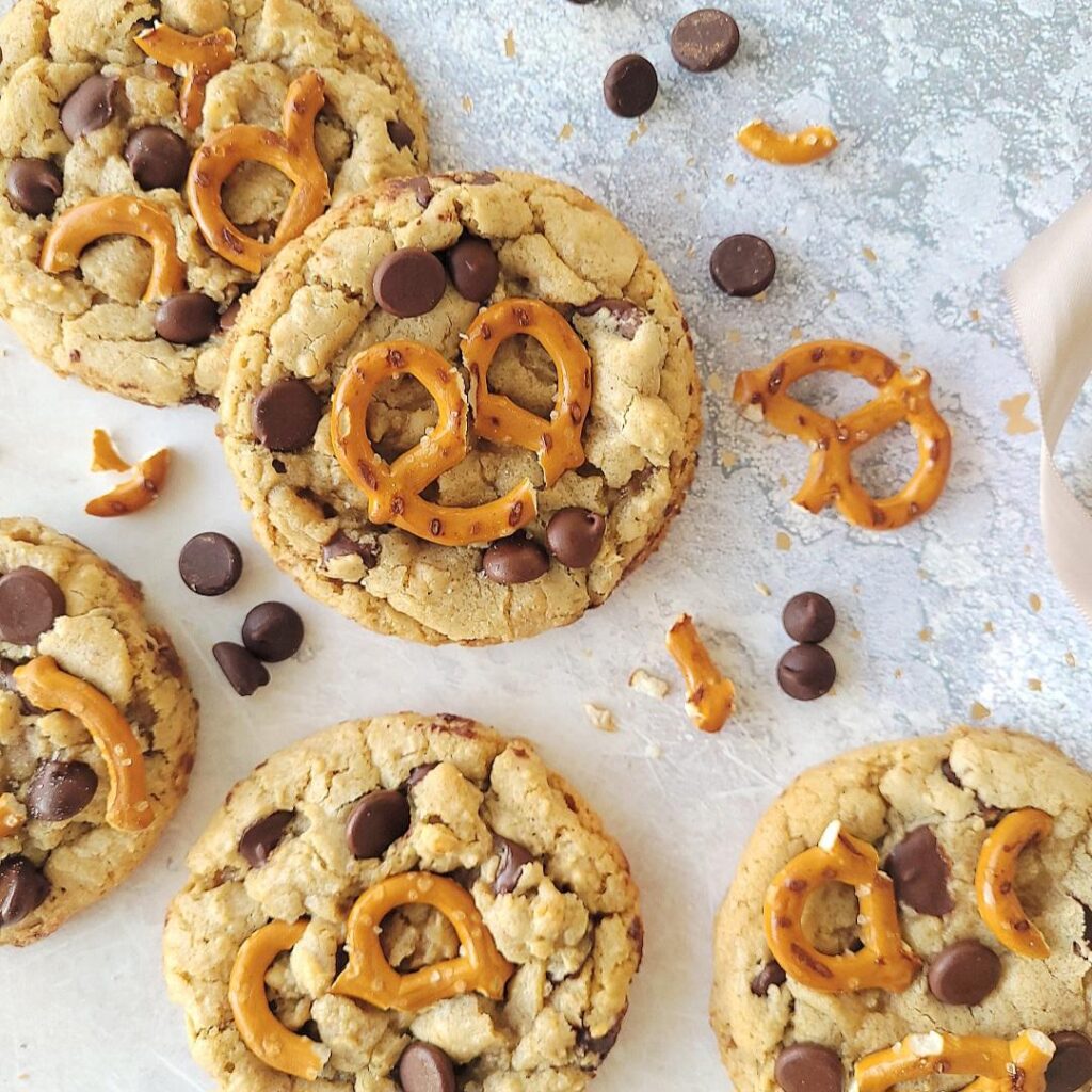 chocolate chip pretzel cookies. top down view of 5 cookies scattered on a gray and white marble surface styled with broken pretzels and loose chocolate chips.