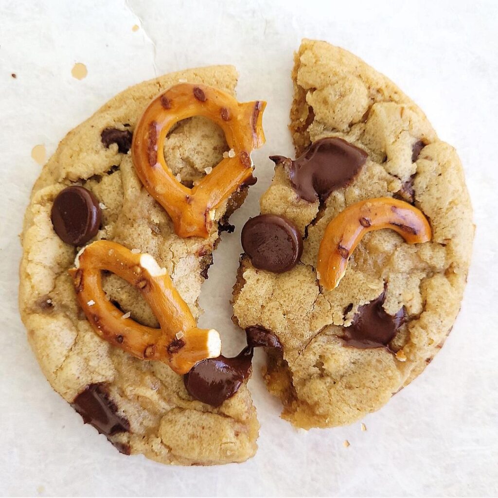 top down view of a single chocolate chip pretzel cookie on a white background. cookie is broken into two halves and topped with pretzel bits and chocolate chips. 