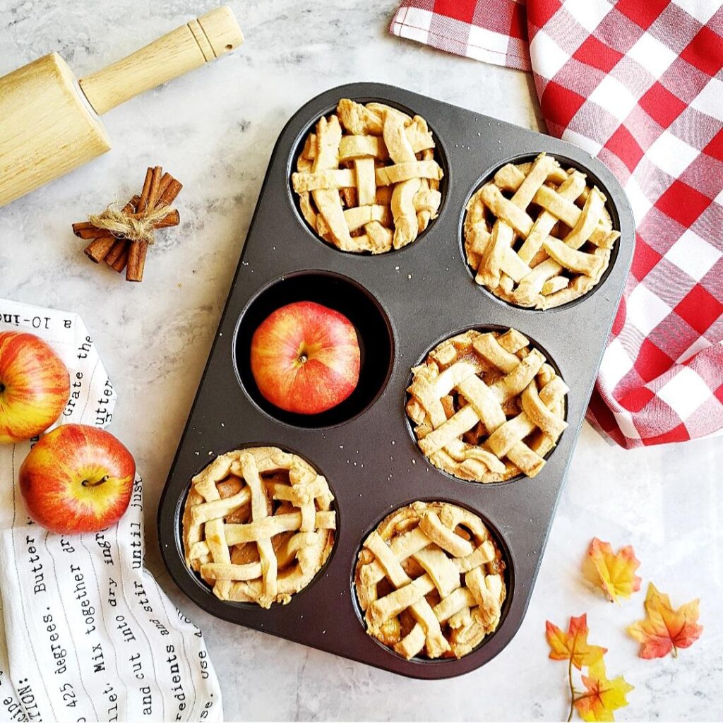 jumbo muffin tin apple pies. top down view of baked apple pies in a jumbo muffin tin pan. one cavity is filled with a red apple instead of a pie. image is styled with apples, fall leaves, cinnamon sticks a rolling pin and a red checkered linen.