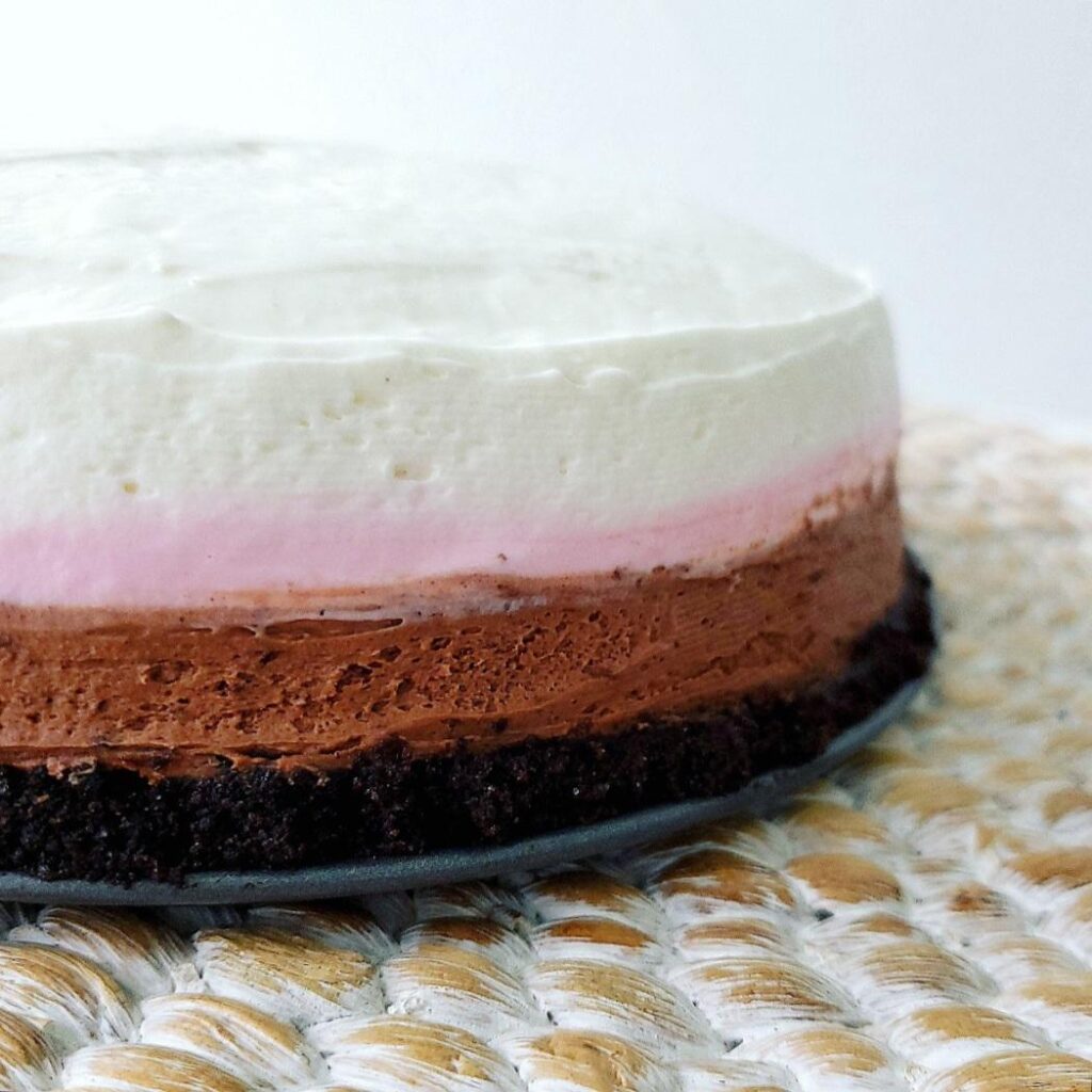 no bake neapolitan cheesecake. side view of cheesecake so you can see the oreo crust on the bottom topped with a chocolate layer, a strawberry layer and a vanilla layer of cheesecake. 
