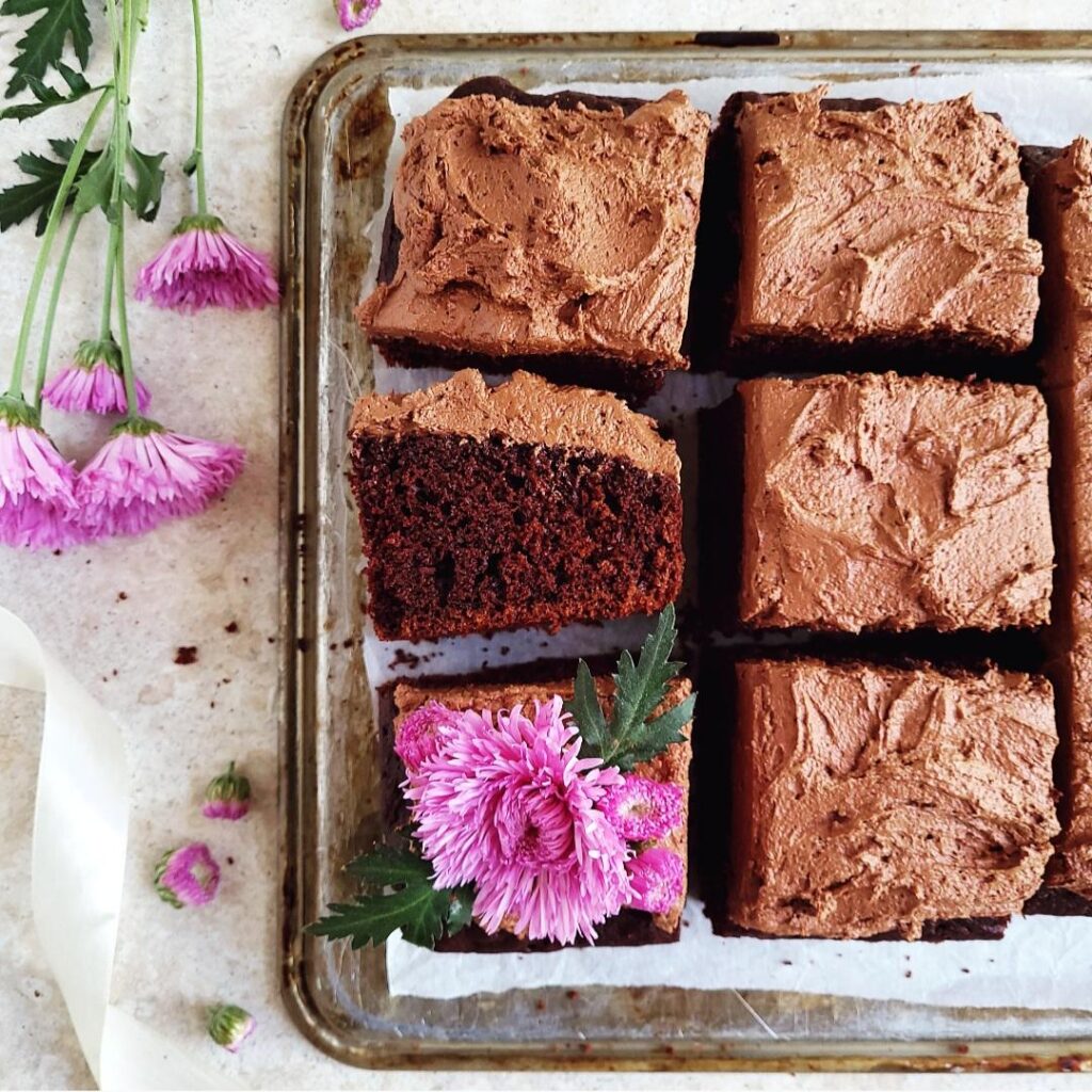 nutella cake with nutella frosting. top down view of the sheet cake cut into squares. one square is flipped on its side so you can see the chocolate cake crumb and thick layer of chocolate frosting. image is styled with purple flowers on the left. 