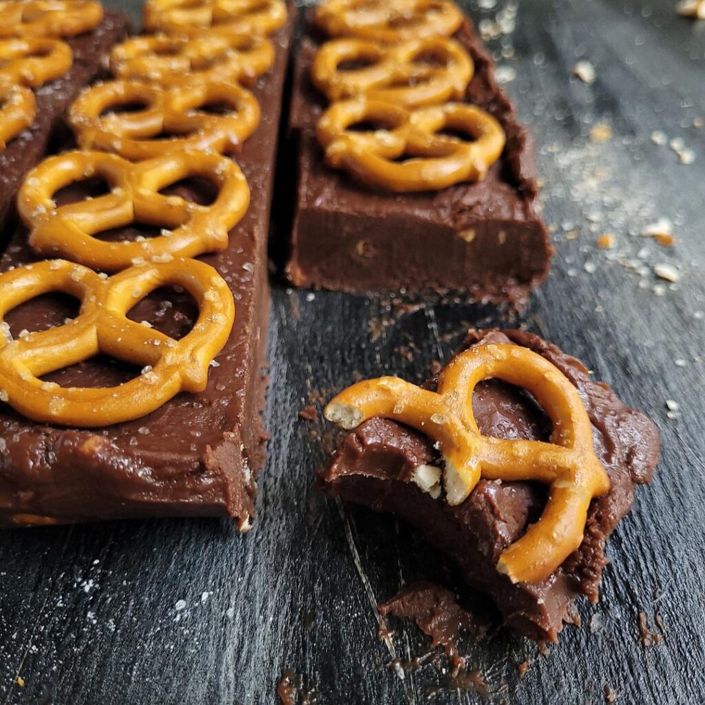 salted pretzel nutella fudge. close up view of a batch of nutella fudge topped with mini pretzels. one square of fudge has been cut away and a bite is missing and the mini pretzel on that slice is broken.  