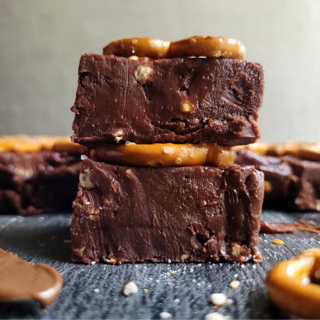 nutella fudge with salted pretzels on top. image is a close up view of two squares of fudge stacked on top of each other. in the background there is a single later of fudge pieces. 