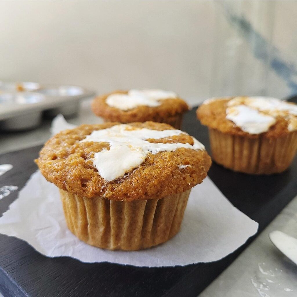 three sweet potato muffins with marshmallow swirled on top. muffins are staggered on a black cutting board. there is a shiny metal muffin pan in the back left corner. 