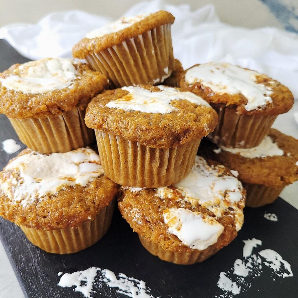 batch of marshmallow swirled sweet potato muffins piled on top of each other. muffins are on a black wooden cutting board splatted with marshmallow fluff. 