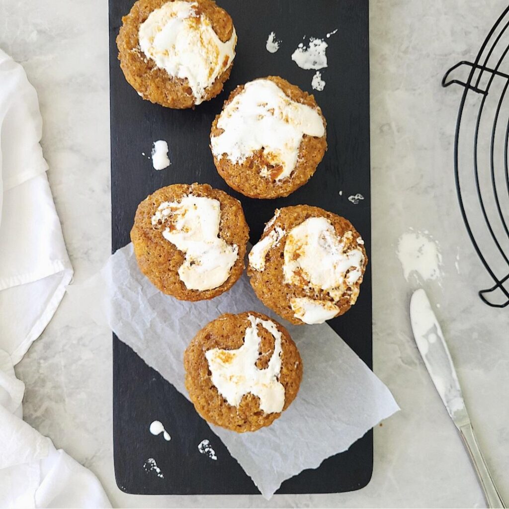 sweet potato muffins with marshmallow fluff on top. top down view of 5 muffins staggered on a black cutting board. there are dips of white marshmallow contrasting on the black wood. background is gray marble. 