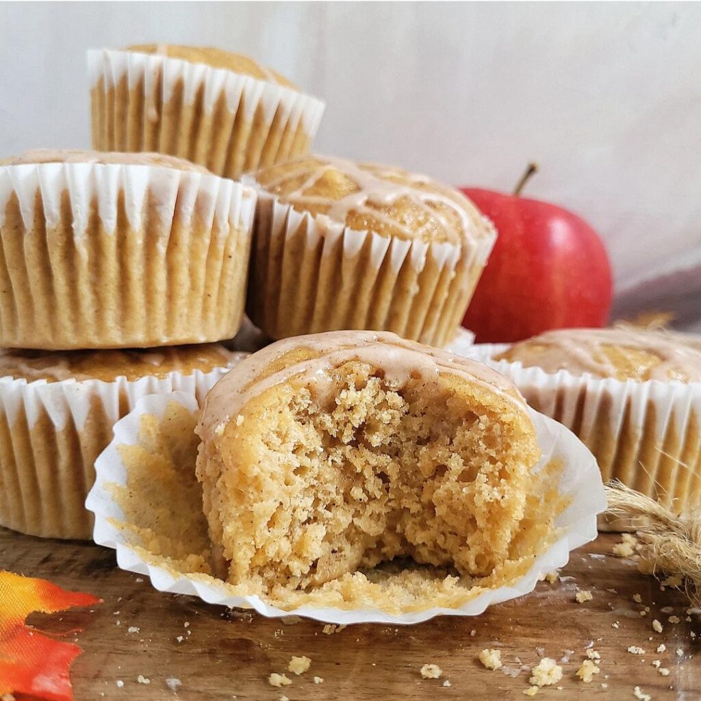 stacked pile of apple cider muffins. muffins have white muffin papers and are dripping with a cinnamon glaze. one muffin has a bite missing so you can see the inner crumb. there is a red apple in the right background. 