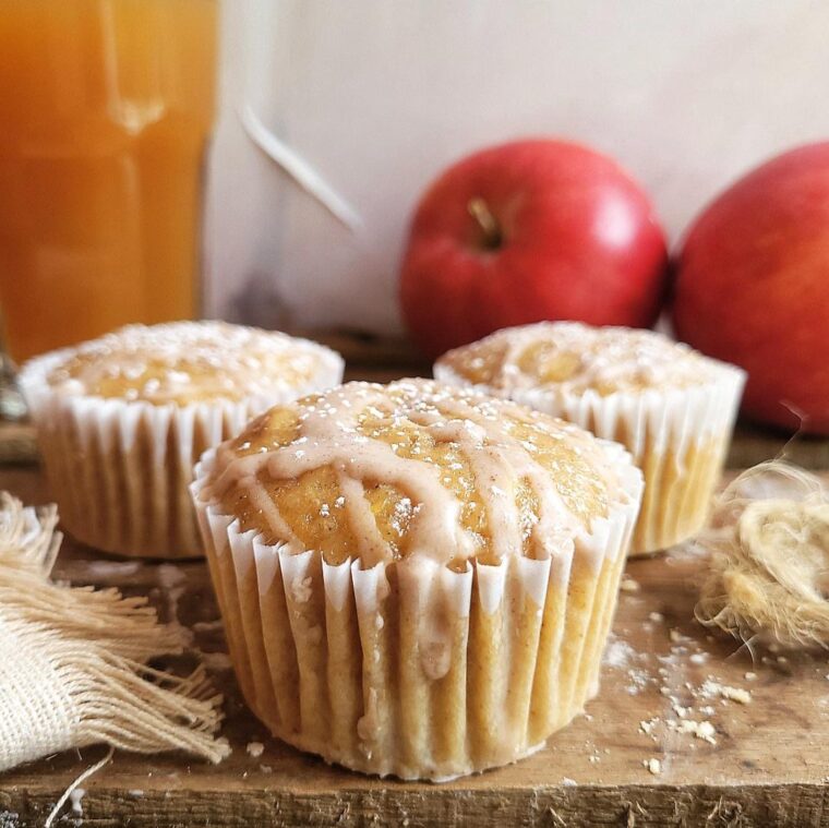 apple cider muffins with cinnamon glaze. side view of three muffins on a rustic wood surface. there are two red apples in the back right and a glass of apple cider in the back left.