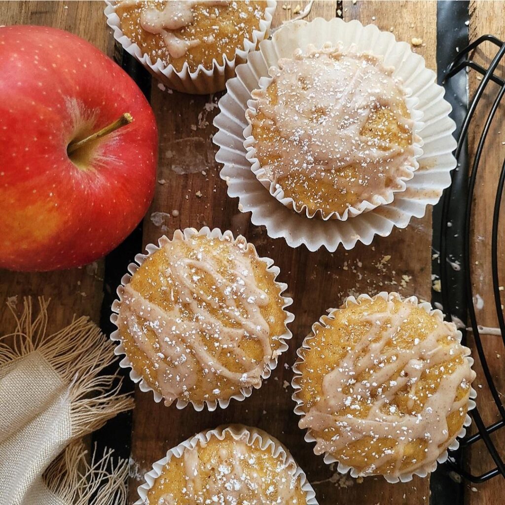 apple cider muffins. top down view of muffins on a rustic wood surface. muffins are dusted with powdered sugar and topped with a cinnamon glaze. there is a red apple on the left. muffins are in white paper muffin liners. 