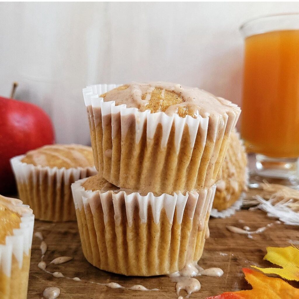 apple cider muffins with cinnamon glaze in white paper muffin liners. two muffins are stacked on top of each other. back left is a red apple, back right is a glass of apple cider. 