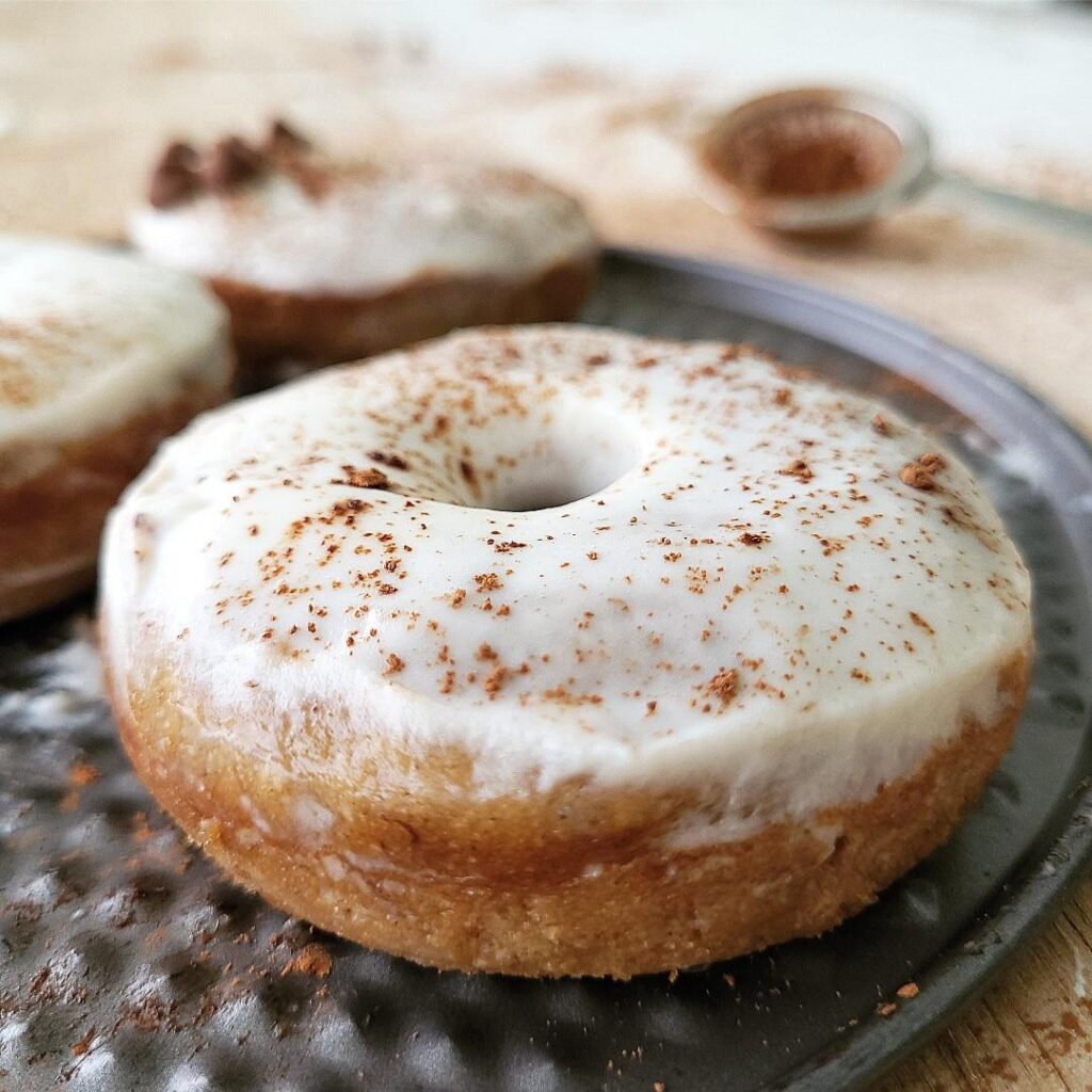 close up 3/4 view of a banana donut with whipped cream frosting dusted with cocoa powder. donut sits on a dark gray charger. there are 2 donuts in the left background.