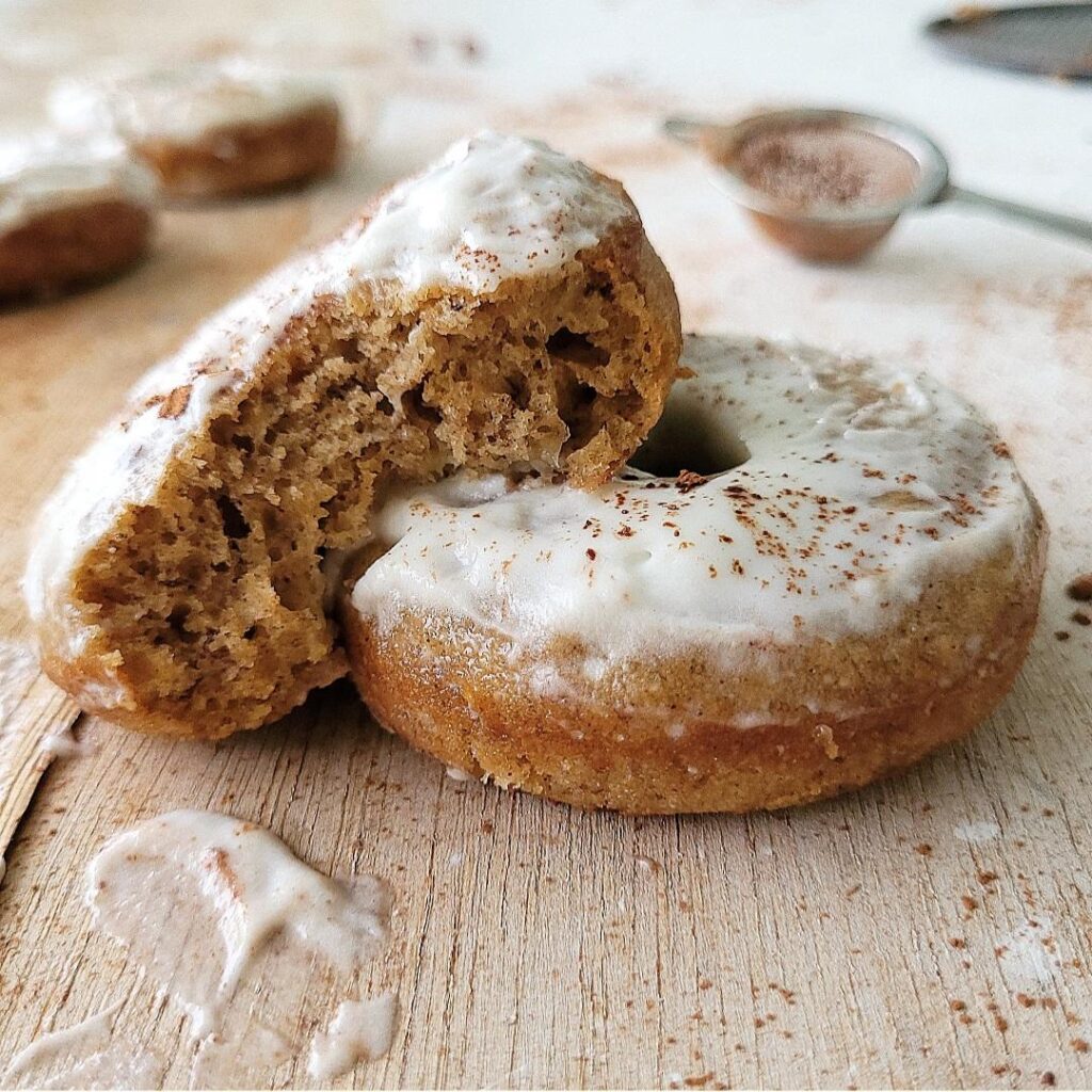baked banana donuts with whipped cream frosting. donuts are dusted with cocoa powder. 3/4 view of a donut on a wood surface with a half eaten donut leaning on it so you can see the inner crumb. 