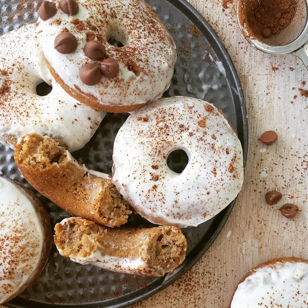 baked banana donuts with whipped cream icing. donuts are dusted with cocoa powder and some are topped with mini chocolate chips. top down view of 5 donuts on a gray charger. A 6th donut is broken in half and both halves are facing up so you can see the inner crumb. 