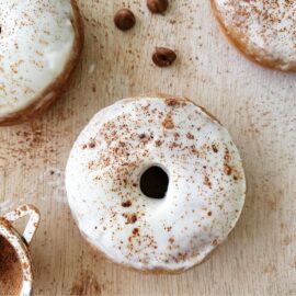 baked banana donuts frosted with whipped cream and dusted with cocoa powder. top down view of a donut on a pale wood surface with partial donuts visible in upper left and right corners.