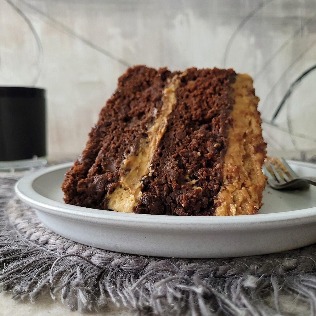 chocolate banana cake with peanut butter frosting. slice of cake on a pale gray plate sitting on a fringed gray fabric charger. background is gray and black abstract. cake is two layers with peanut butter frosting in the middle and on top. 