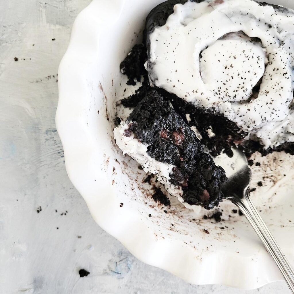 top down view of a chocolate cinnamon roll with white icing in a white bowl. background is pale gray with some black crumbs. a forkful of the cinnamon roll has been pulled away and turned on its side so you can see the chocolate crumb and chocolate chips inside.  