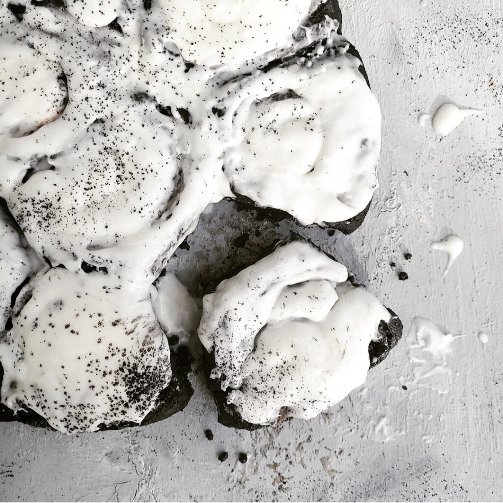 chocolate cinnamon rolls with white icing sprinkled with black cocoa powder. top down view of rolls on a pale gray background. one roll has been cut from the rest and is askew and surrounded by black crumb and drops of white icing. 