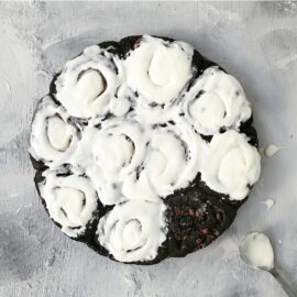 chocolate cinnamon rolls. top down view of uncut rolls. all but one has been frosted with white icing. background is pale gray and image is styled with a metal spoon with icing on it.