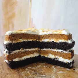 four later chocolate pumpkin cake with cream cheese frosting. side view of cut cake on a wooden surface. slices have been removed from cake so you can see layers of pumpkin cake and cream cheese frosting stacked on layers of chocolate cake.