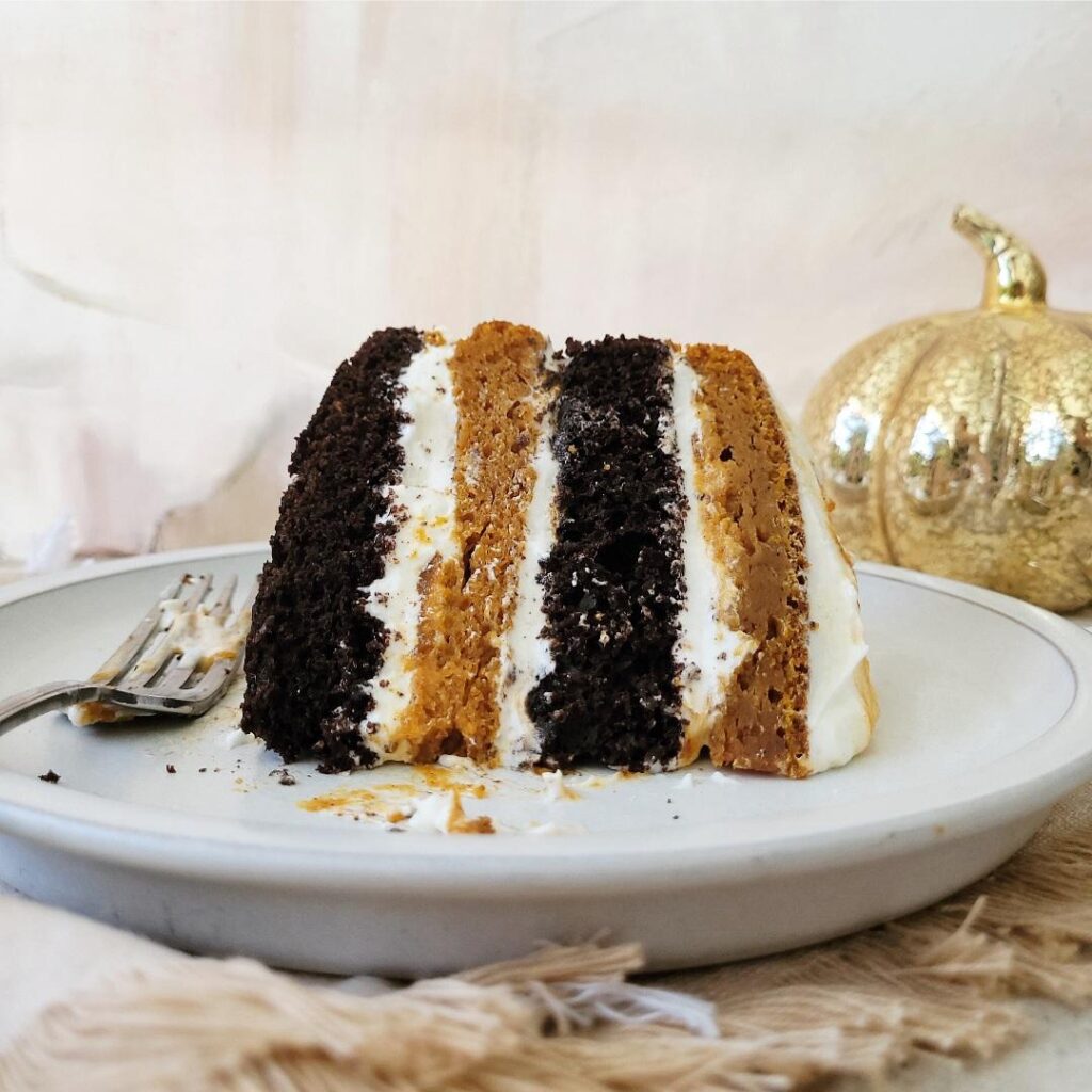 four layer chocolate pumpkin cake with cream cheese frosting. slice of cake laying on a pale gray plate with a gold pumpkin decoration in the back right corner. 