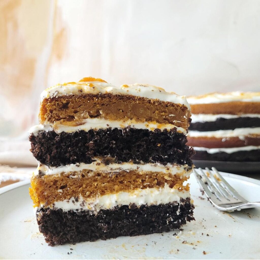 four layer chocolate pumpkin cake slice standing tall on a plate with the remaining uncut cake in the back right corner. cake has alternating black chocolate and orange pumpkin layers with white cream cheese frosting in between layers. 