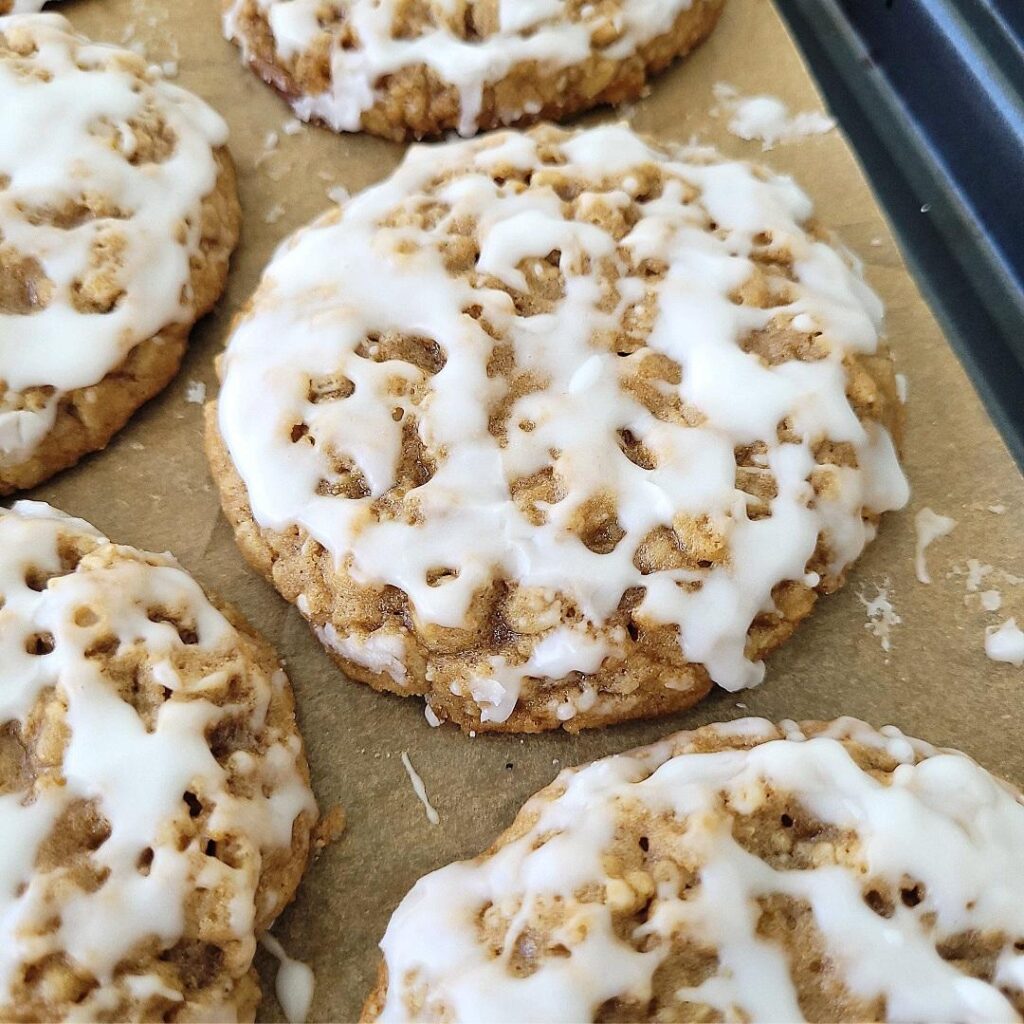 old fashioned iced oatmeal cookies on a dark colored metal baking pan lined with brown parchment that has drips of vanilla glaze 