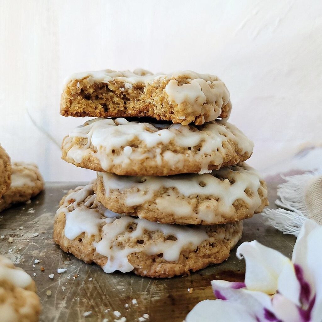 chewy oatmeal cookies iced with a vanilla glaze. side view of 4 cookies stacked on top of each other. the top cookie has been bitten so you can see the inner crumb. background is abstract white with texture. 