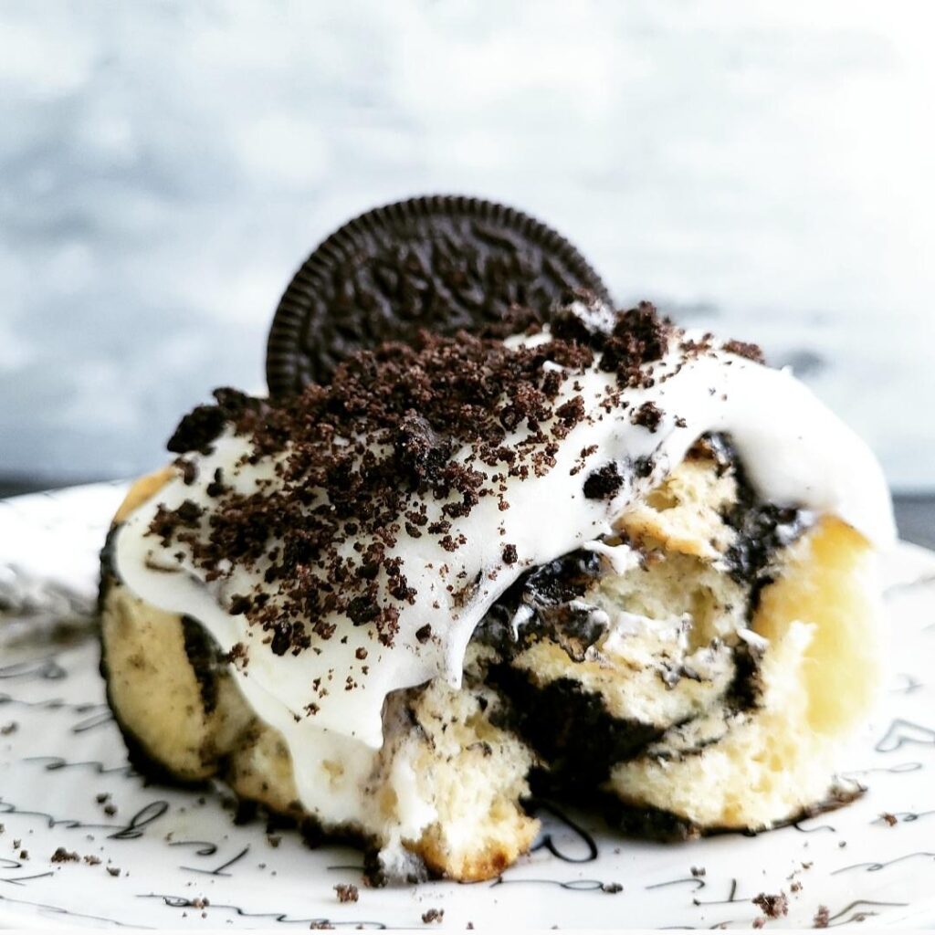 oreo cinnamon rolls. image of one cinnamon roll topped with cream cheese frosting, oreo cookie crumbs and a horizontal oreo cookie. side view of cinnamon roll so you can see the inner swirl of black cocoa in the soft fluffy dough. 