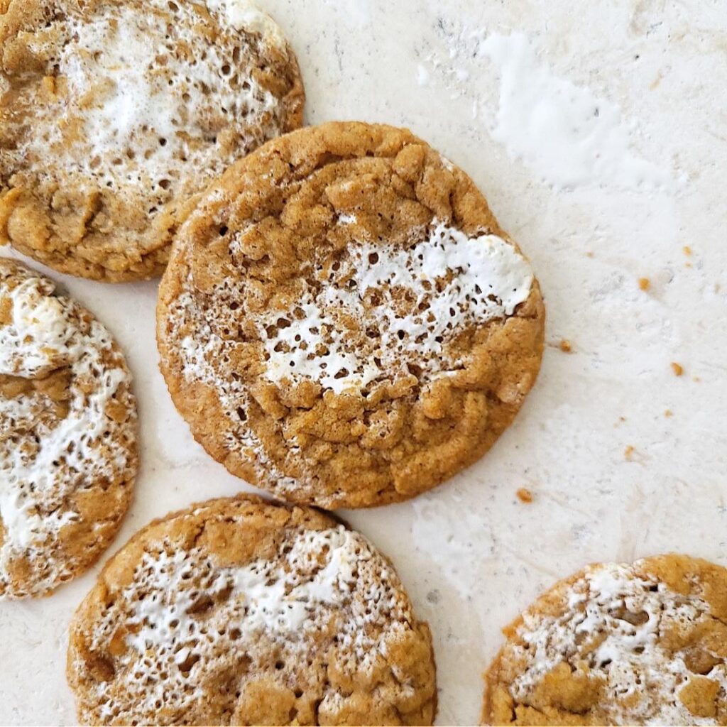 pumpkin marshmallow cookies. top down view of five cookies scattered on a white background with a few cookie crumbs. cookies are pumpkin colored with swirls of marshmallow fluff. 
